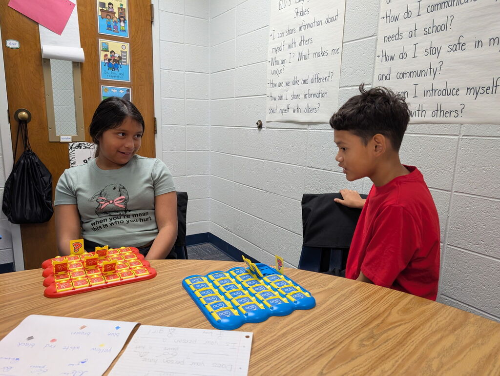 Two students, a girl and a boy, sit at a table playing a board game. Papers and a bag hang in the background.