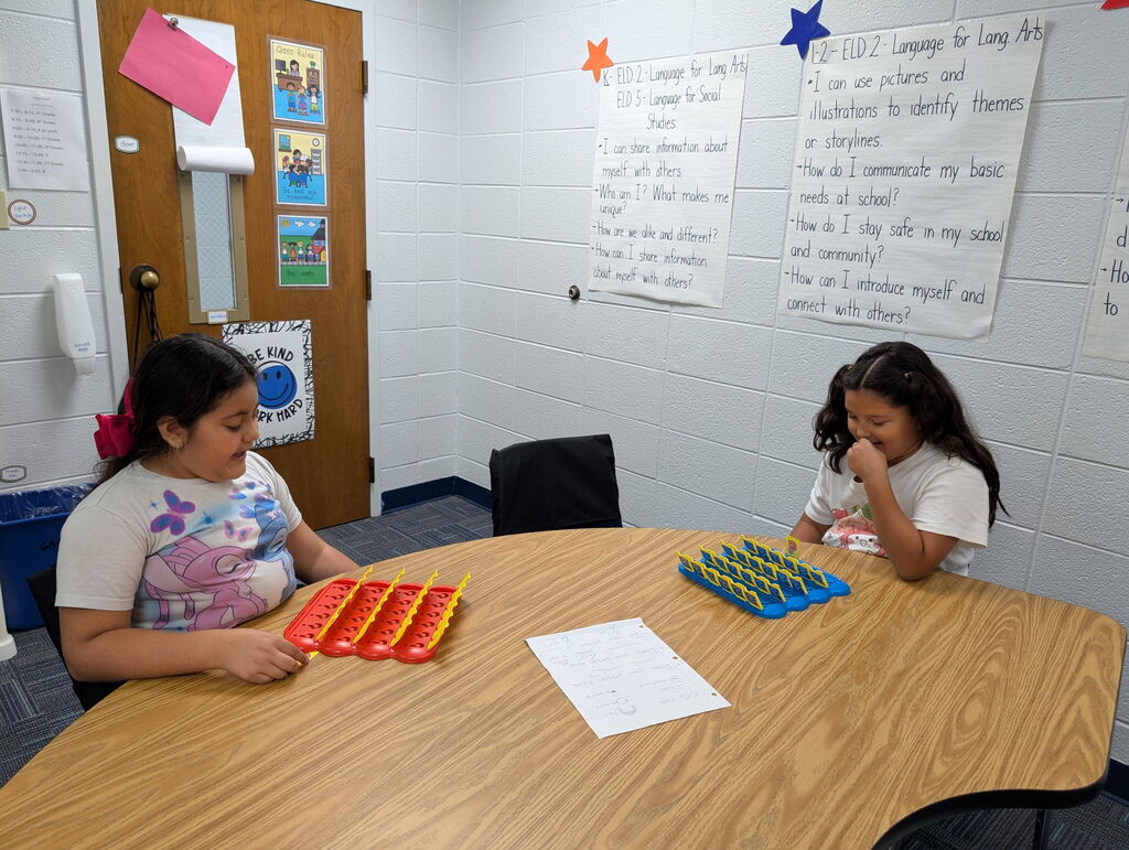 Two children play a board game on a wooden table in a classroom with white walls and bulletin boards.
