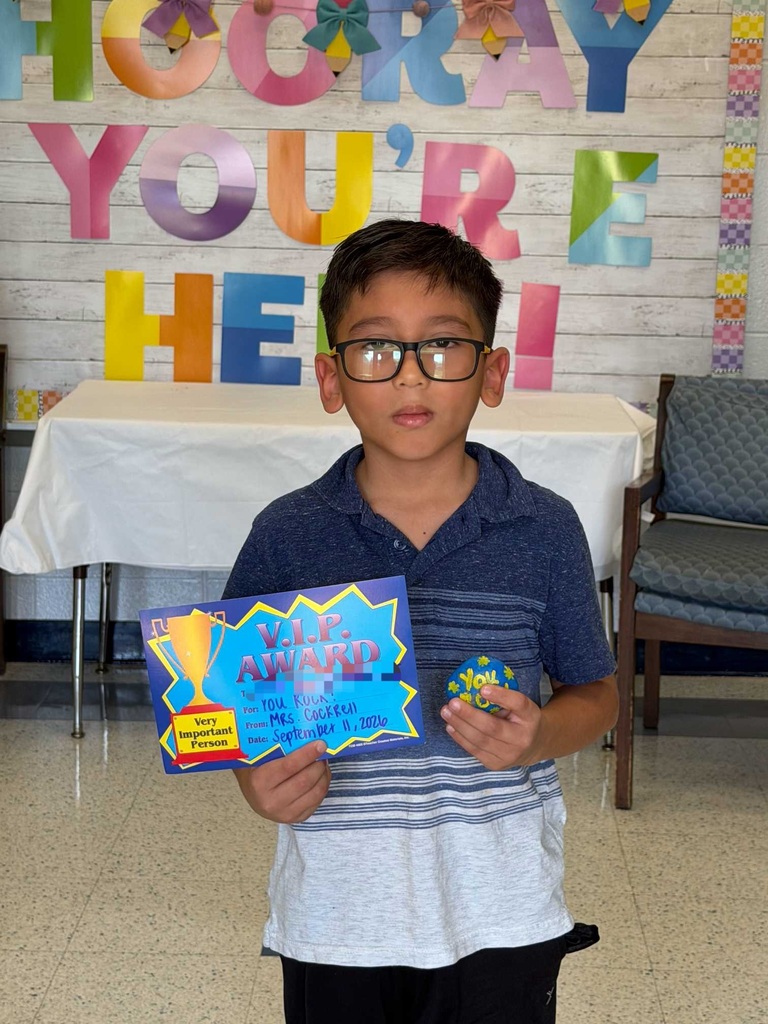 A young boy holds a colorful award certificate and a small round object. Behind him is a desk with a white cloth and a wall with colorful letters.