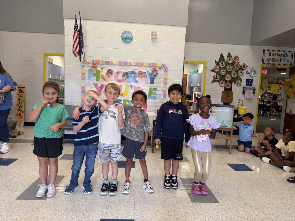 Children stand together in a classroom, smiling, with a banner behind them. Some are wearing sneakers and one wears pink shoes.