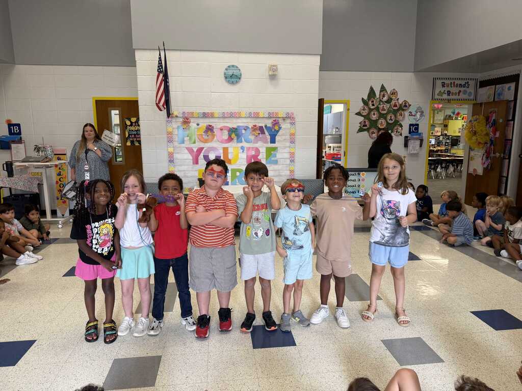 Nine children stand in a row in a classroom, wearing various clothing and footwear. A woman stands near a flag in the background.