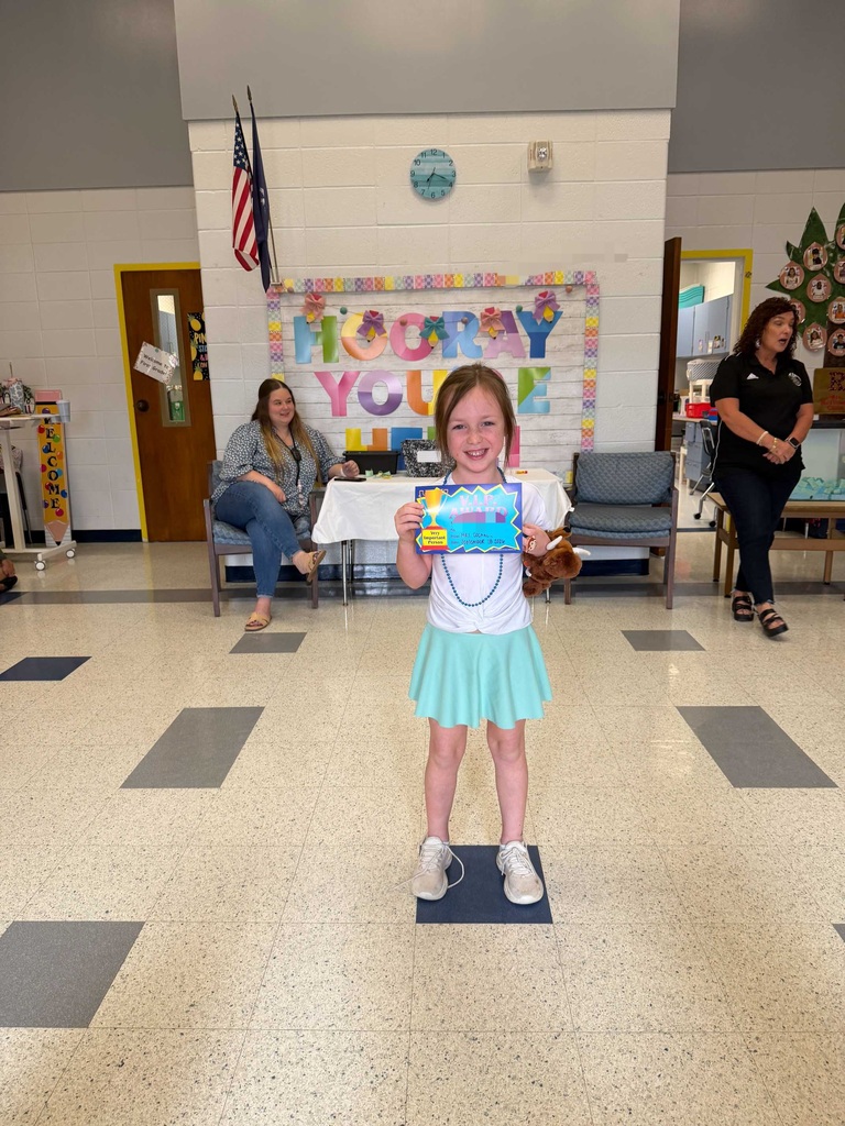A young girl stands on a mat, holding a book and smiling. Behind her, people sit and stand, with a table, chairs, and a bulletin board in a school hallway.