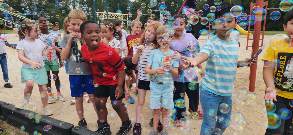 A group of children outdoors playing with bubbles. They are standing in a dirt area with some playground equipment.