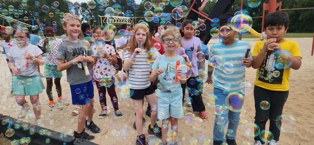 A group of children standing in sand play with bubble wands. Some bubbles float in the air.