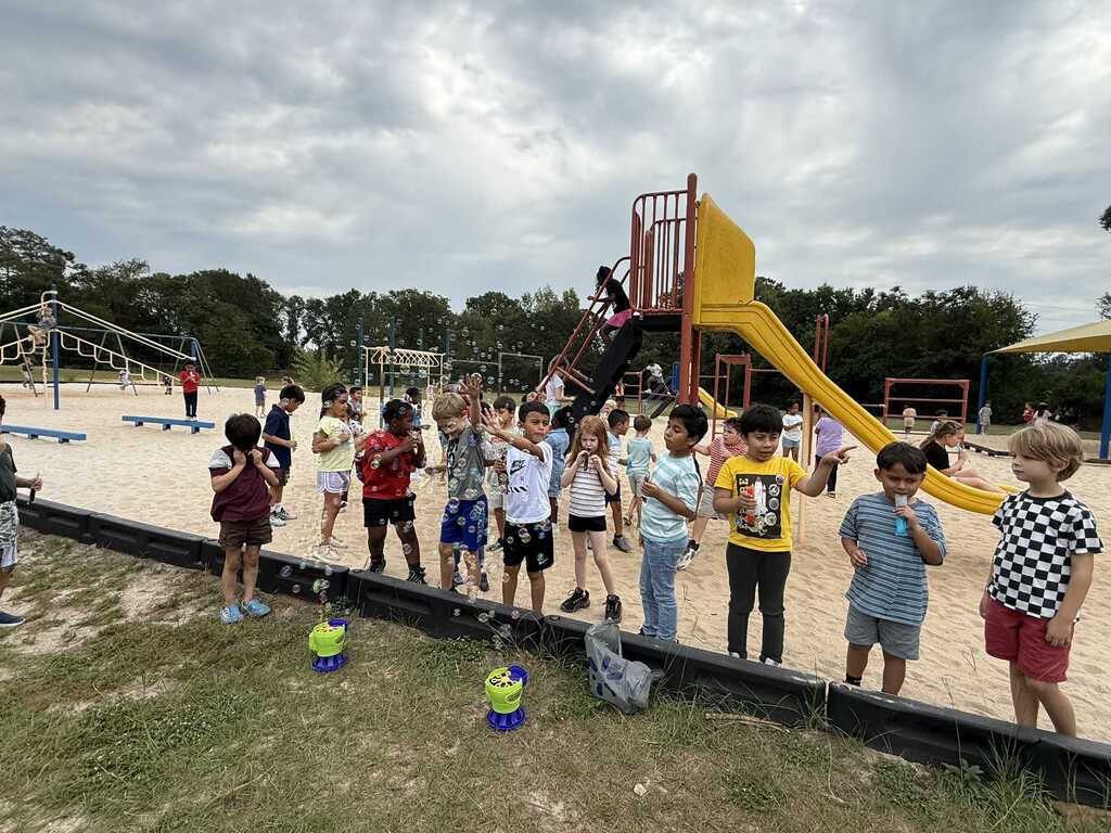Kids stand in a playground. One child holds a bubble maker. Behind them is a slide, a climbing frame, and other playground equipment.