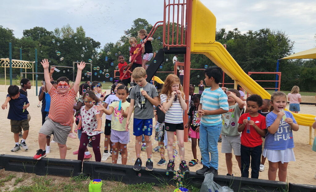 A group of children and adults are at a playground. They are standing and some are sitting on the yellow slide.