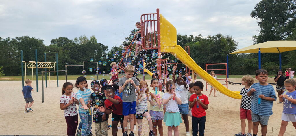 A group of children in a park with a yellow slide and a bubble machine. Some children are holding bubbles.