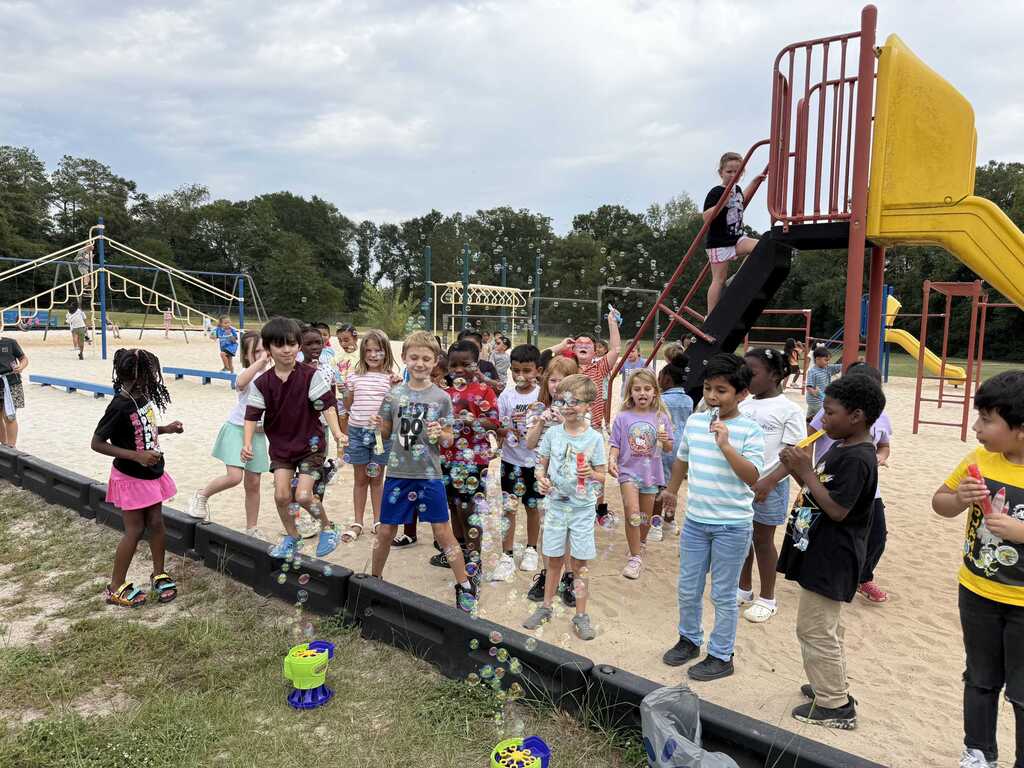 Children gather near playground equipment at a park, some holding bubble wands. A yellow slide is in the background.