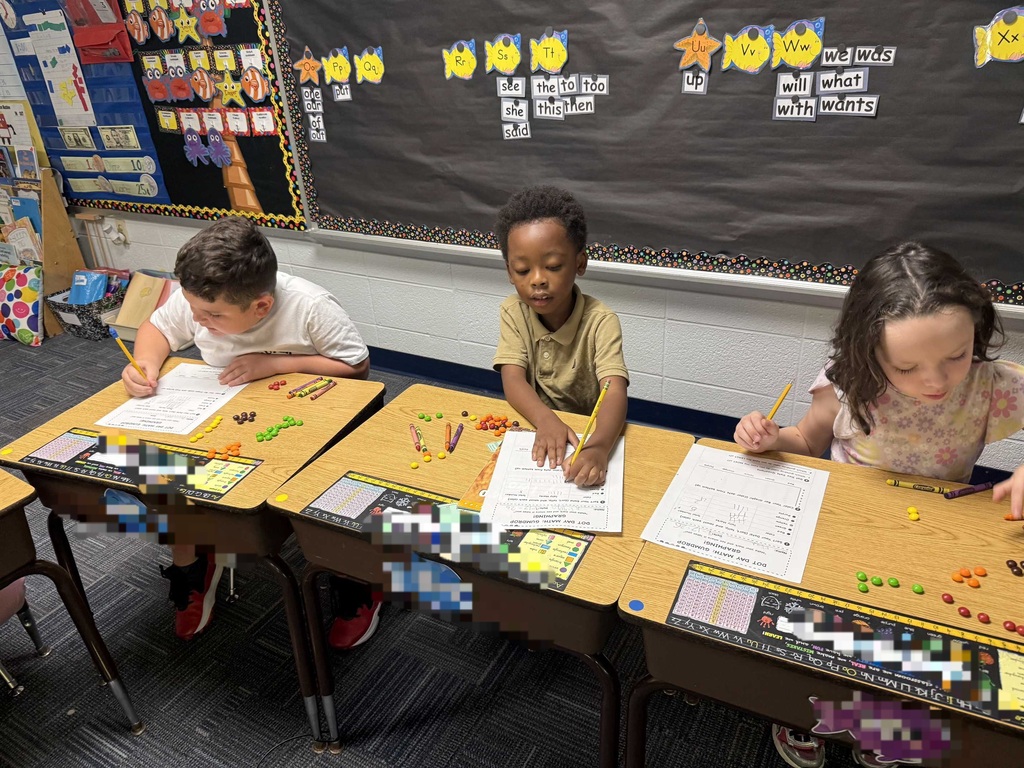 Three children at desks in a classroom with a bulletin board behind them. They have papers and colorful objects on the desks.
