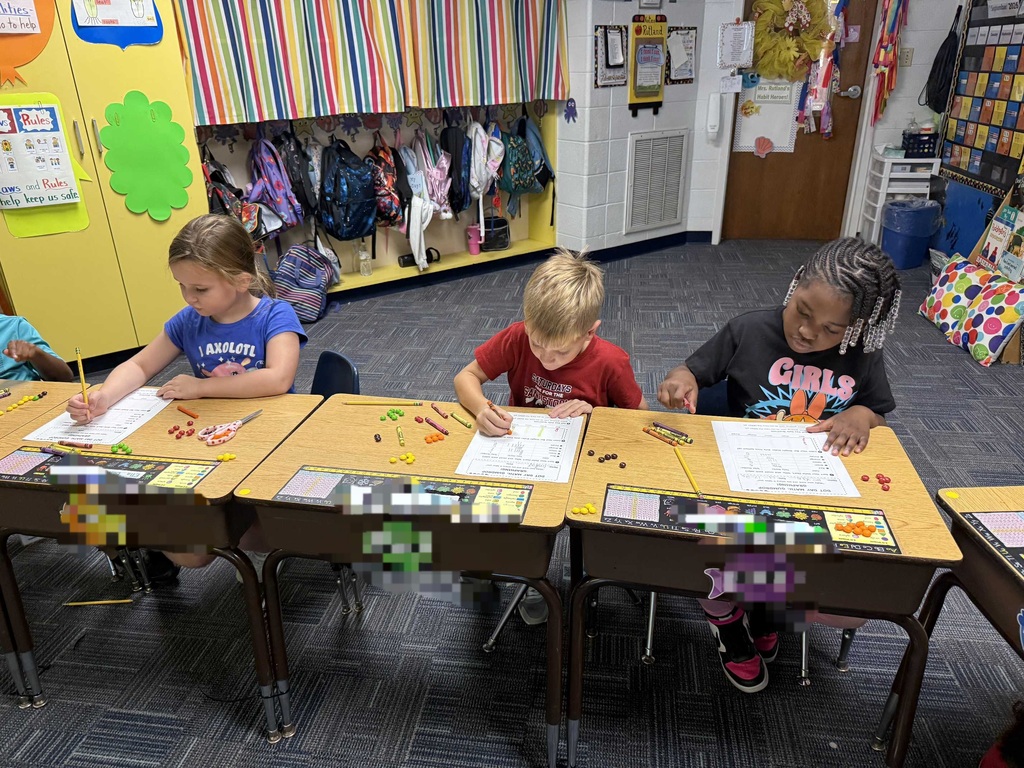 Four children sitting at desks, doing activities. One holding a marker. Background shows a classroom with colorful wall decorations.