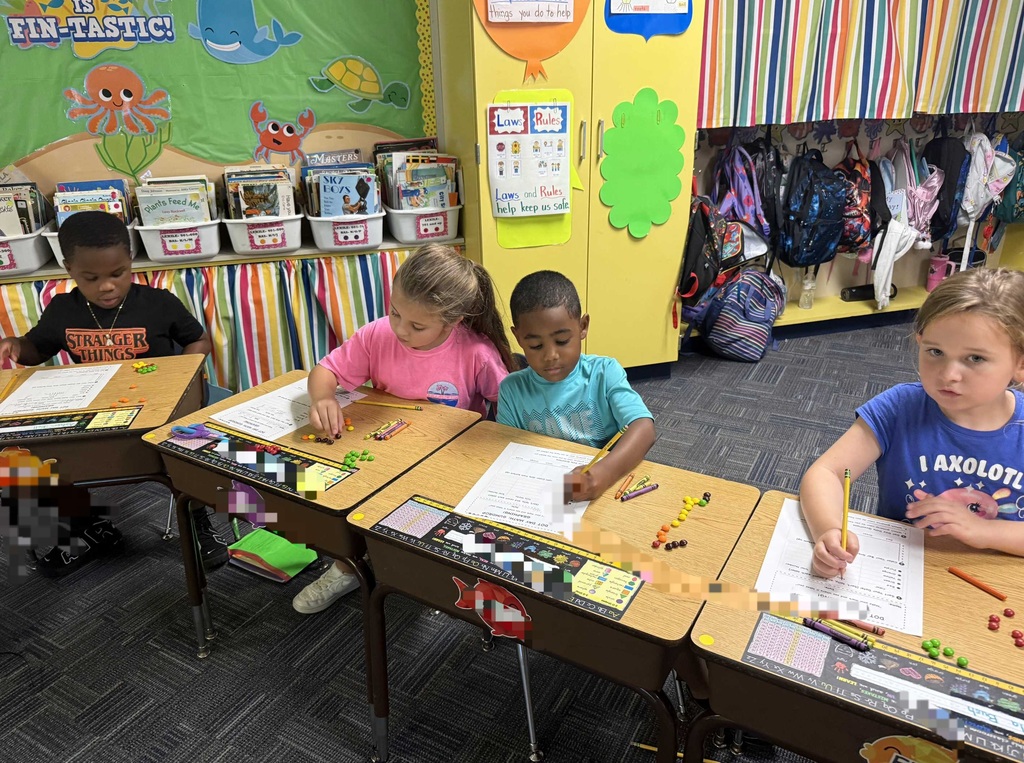 Four children seated at desks with papers and crayons, in a classroom with colorful decorations and shelves.