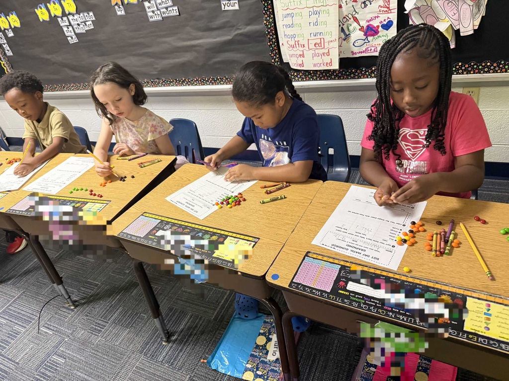 Four children are seated in a classroom. They work at desks with papers, crayons, and various colorful objects.