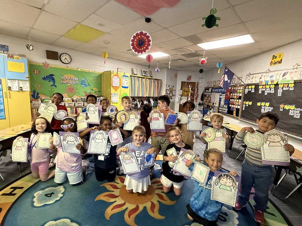 Classroom with children holding up certificates; tables, chairs, and decorations in background.