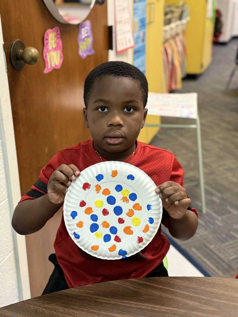 Child wearing a red shirt holding a paper plate with multicolored dots in a classroom.