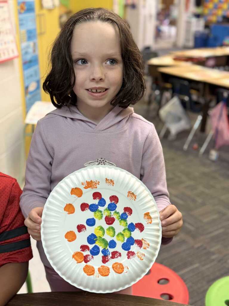 A child holds a paper plate with colorful dots, standing in a classroom with tables and chairs.