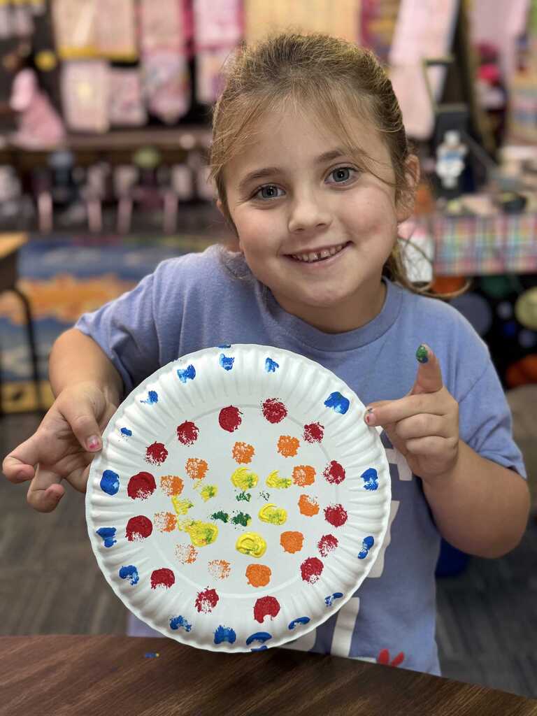A girl in a blue shirt holds a paper plate with colorful dots. She is smiling.