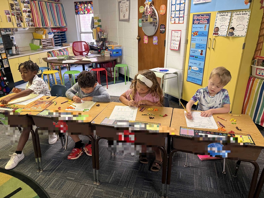 Four children sit at desks, working on crafts, with a colorful classroom background. Brightly colored classroom decorations are visible.