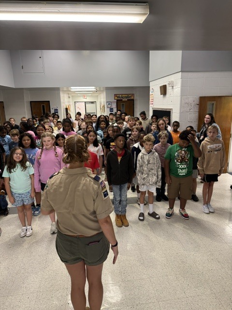 People gather in a hallway. A woman stands at the front, addressing the group. Some wear casual clothing.