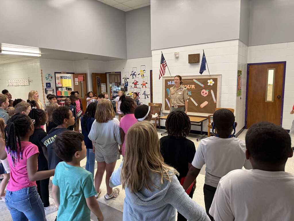 In a classroom, a group of children listens to a woman in a military uniform standing near a bulletin board.