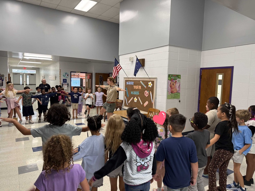 A group of children forms a circle around an adult in a school hallway. The adult holds a flag.
