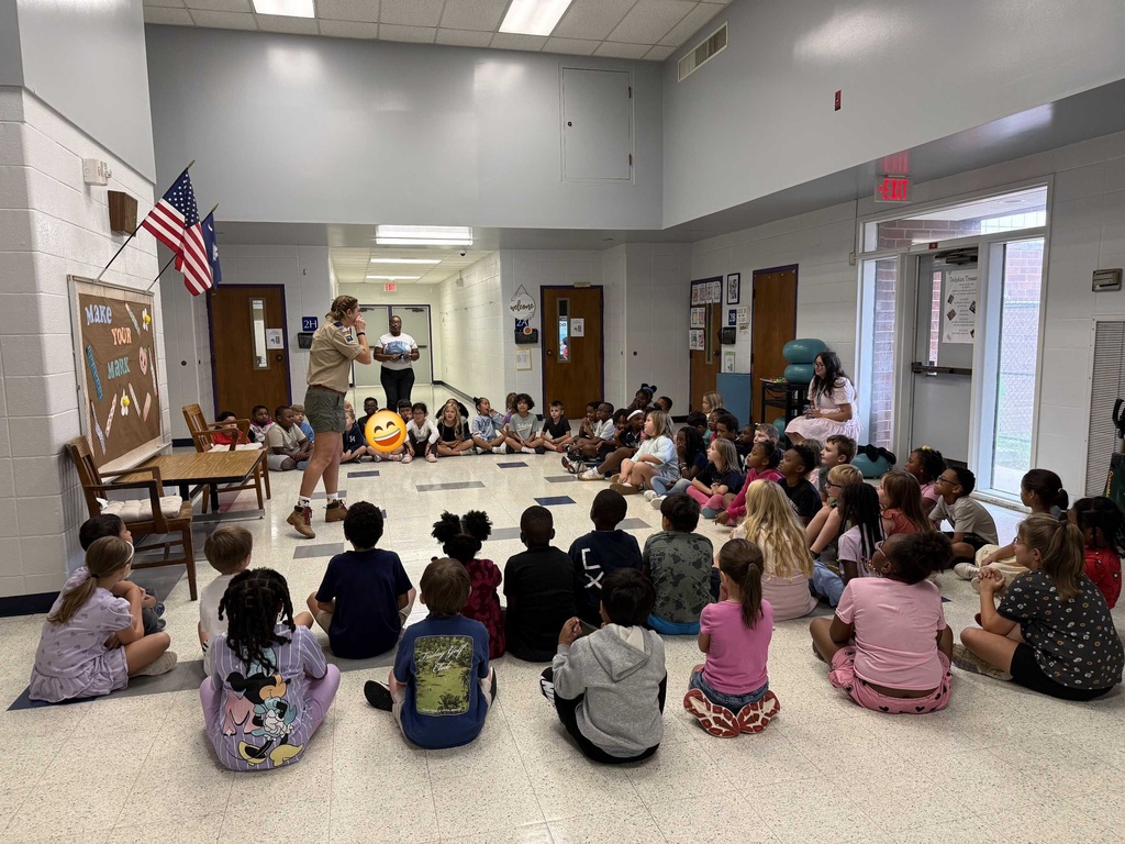 A classroom with kids seated in a circle; a woman stands in front, an American flag hangs on the wall.
