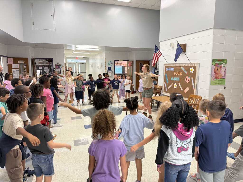 A classroom filled with children standing in a circle around an adult, with American flags and bulletin boards in the background.