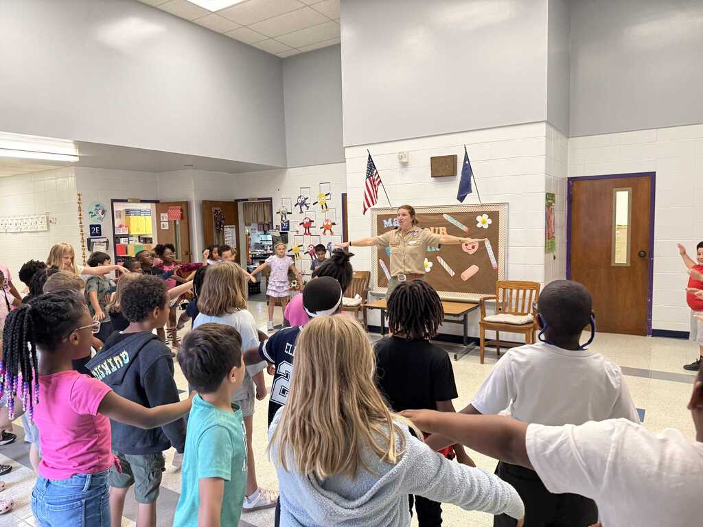 A group of children in a classroom form a circle around a woman, possibly their teacher, who holds objects.
