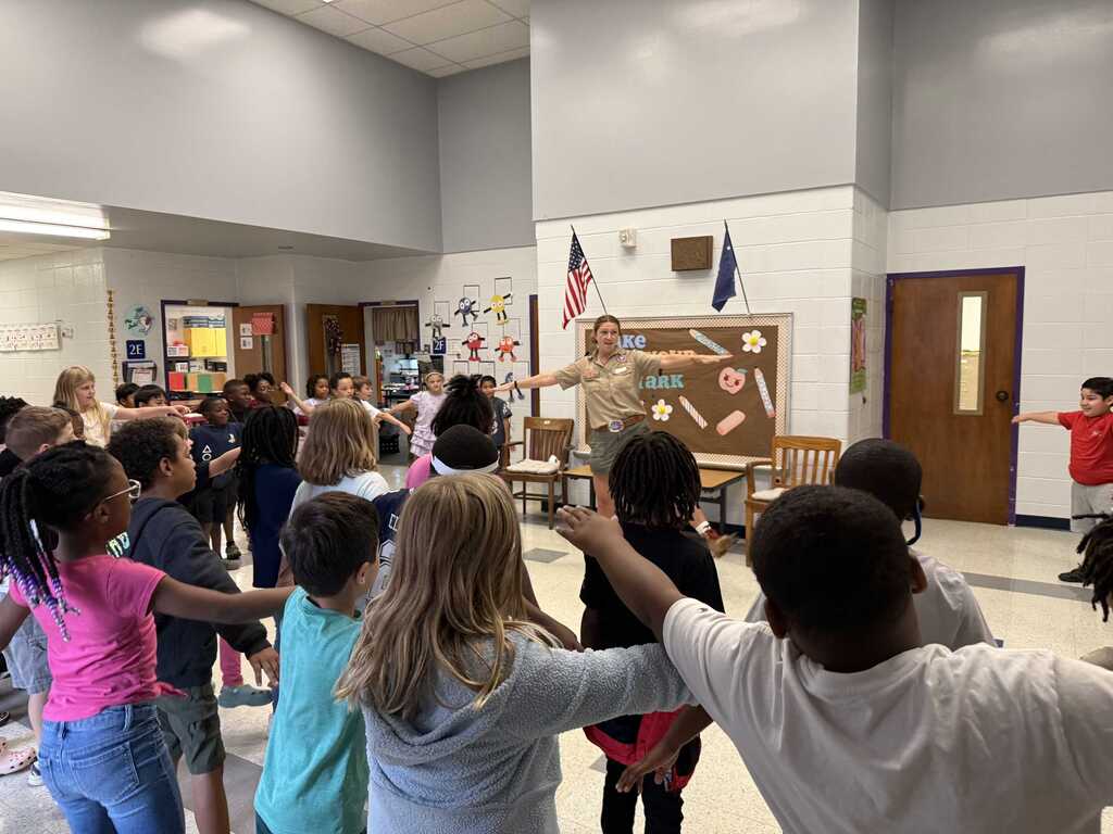 A classroom with children holding hands in a circle. A uniformed adult stands in the center.