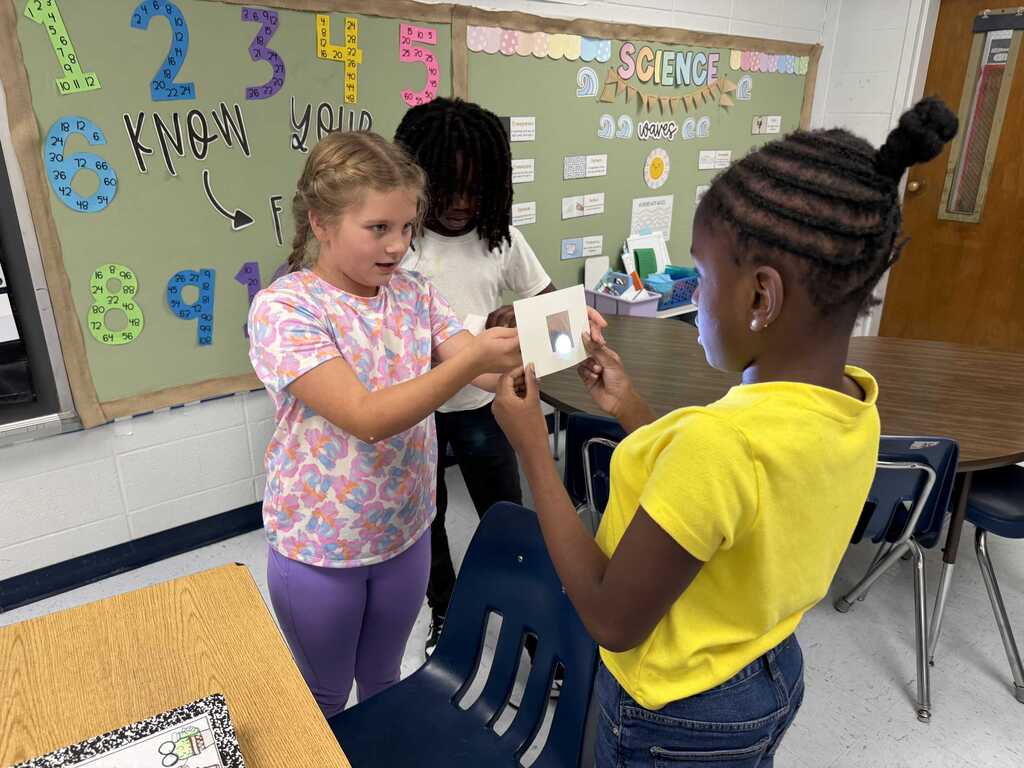 Three students in a classroom. A girl hands another a small card. Behind them is a green bulletin board with letters and numbers.