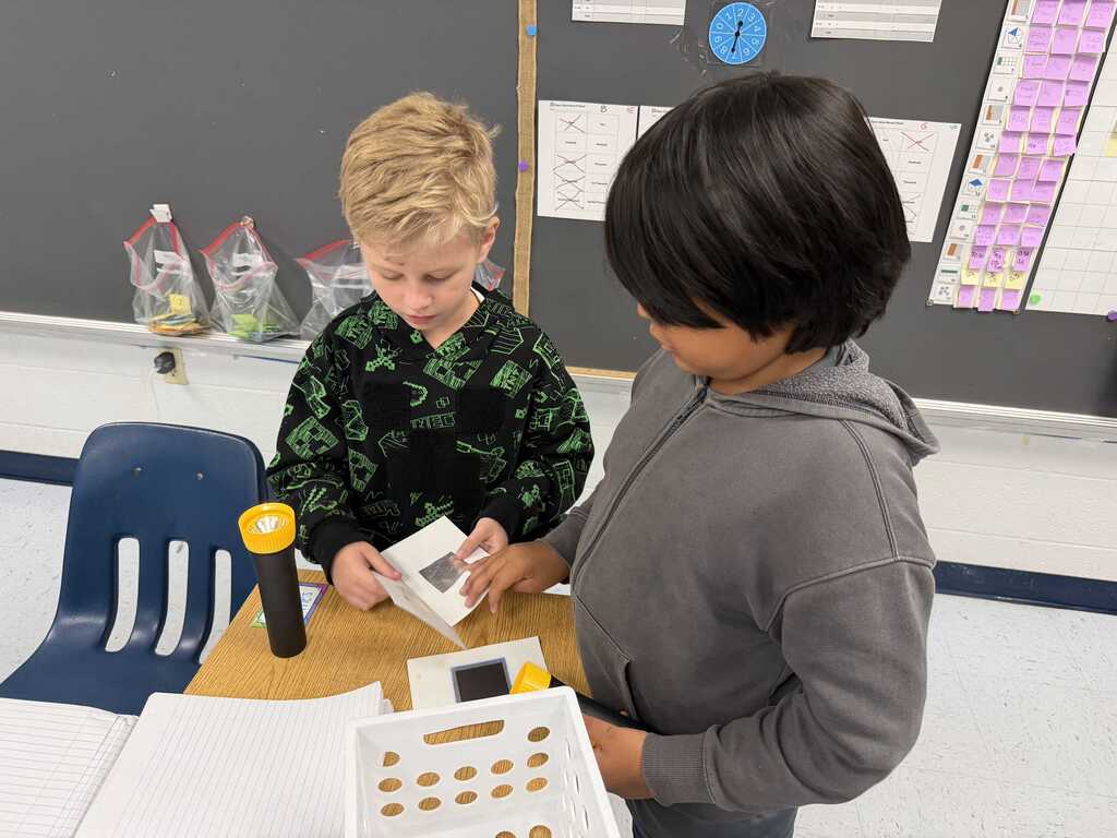 Two children in a classroom, one looking at a box, the other holding a box. A desk is between them.
