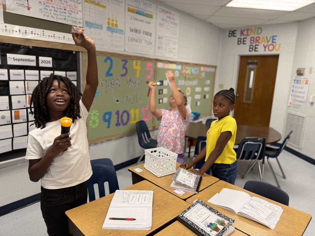 Three children stand in a classroom, one holding a microphone, another holding a pointer, and a third reading from a book.