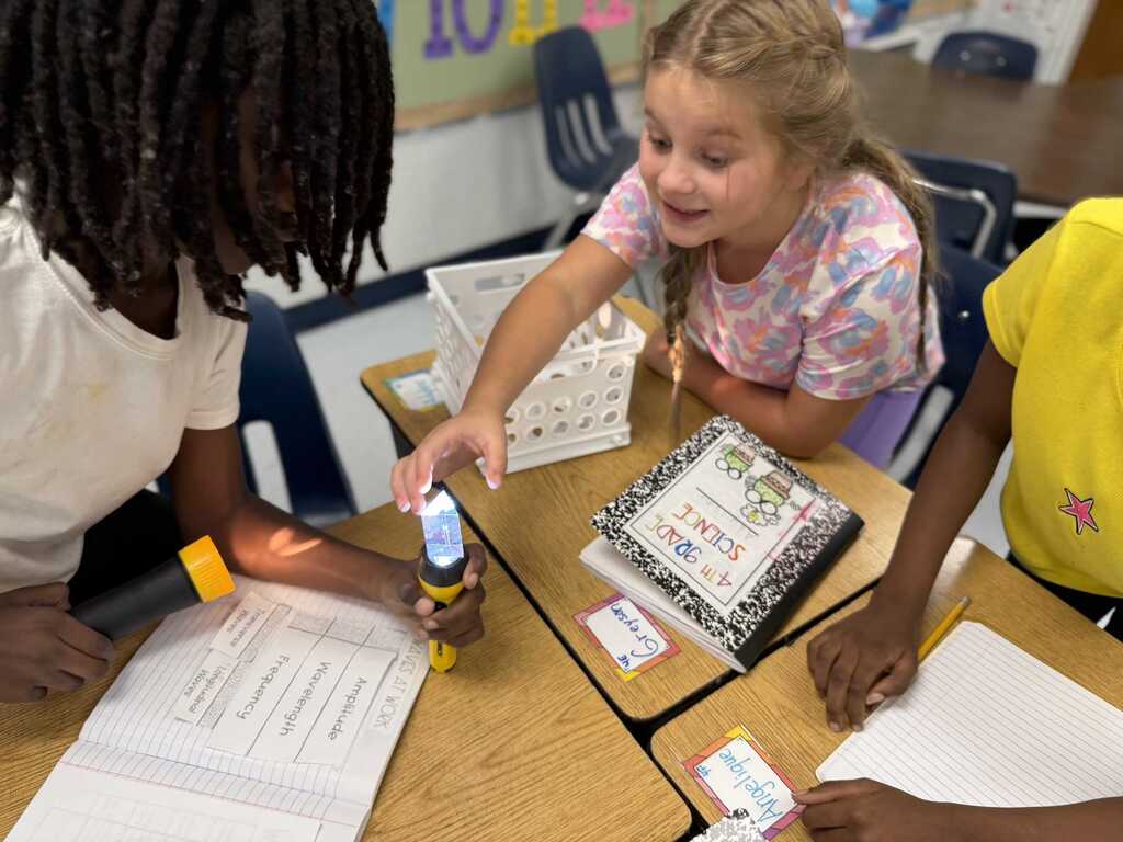 Three children at desks, one holding a flashlight, another with a book, and the third with a pencil.