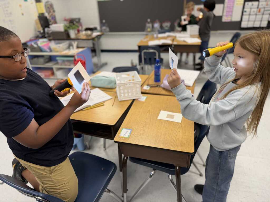 Two students are in a classroom, one using a magnifying glass and the other holding papers.