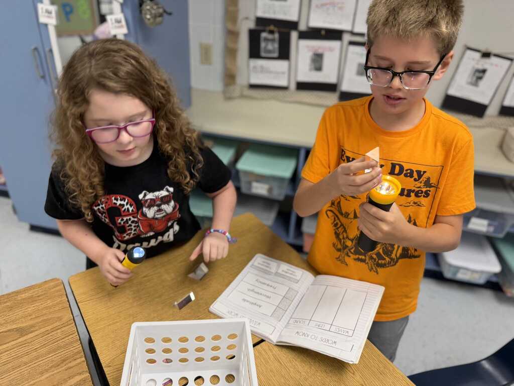 Two kids, one in orange and the other in black, work on a project at a desk with a white tray, papers, and small objects.