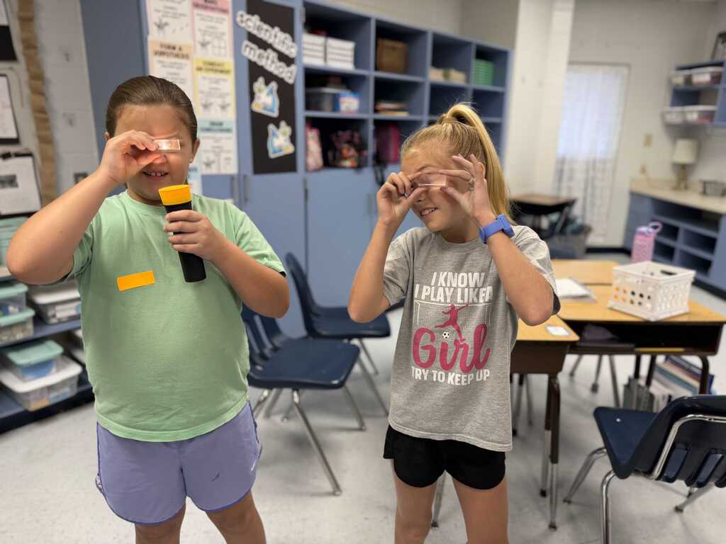Two girls are standing in a classroom, each holding a magnifying glass. They appear to be focused on something. Behind them are chairs, a desk, and storage shelves.