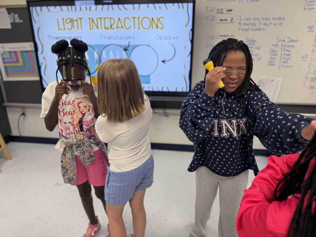 Four students in a classroom use yellow flashlights to demonstrate light interactions. A large screen displays related information.