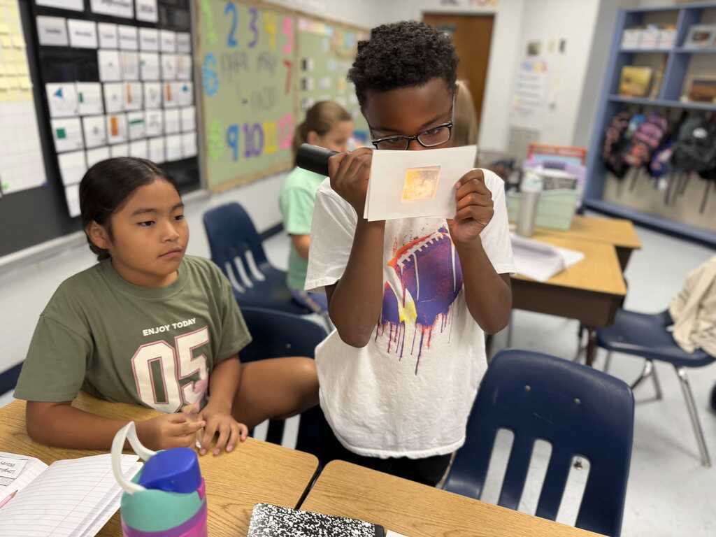 Two students in a classroom hold up a paper with a yellow circle, one wearing a green shirt.