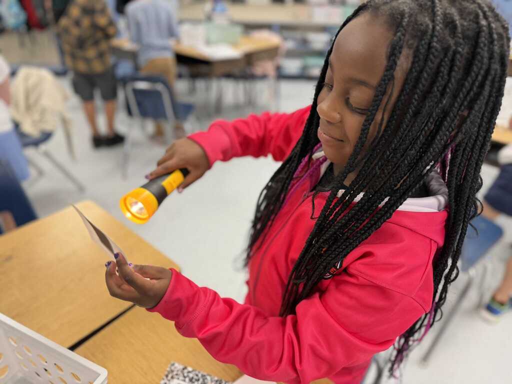 A girl with braided hair uses a flashlight to examine a small object on a desk.