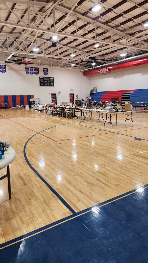 An empty indoor basketball court with blue lines and hardwood floors. Tables with items sit near the center.