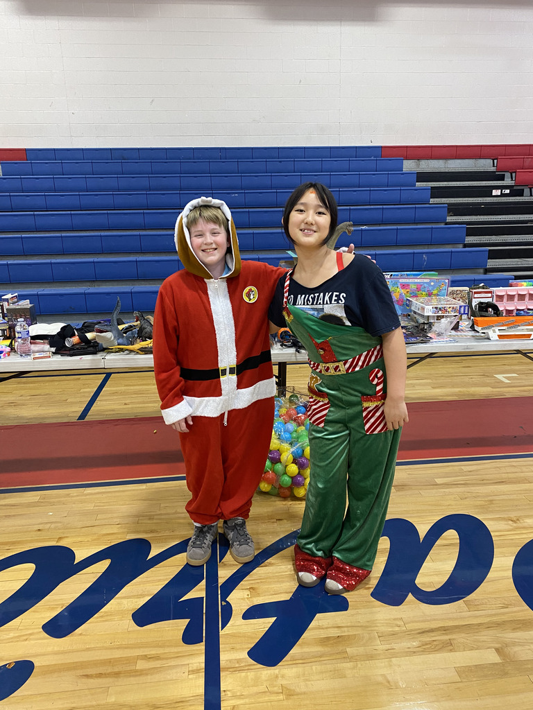 Two people stand on a basketball court. One wears a Santa Claus costume, and the other wears a Christmas jumpsuit.