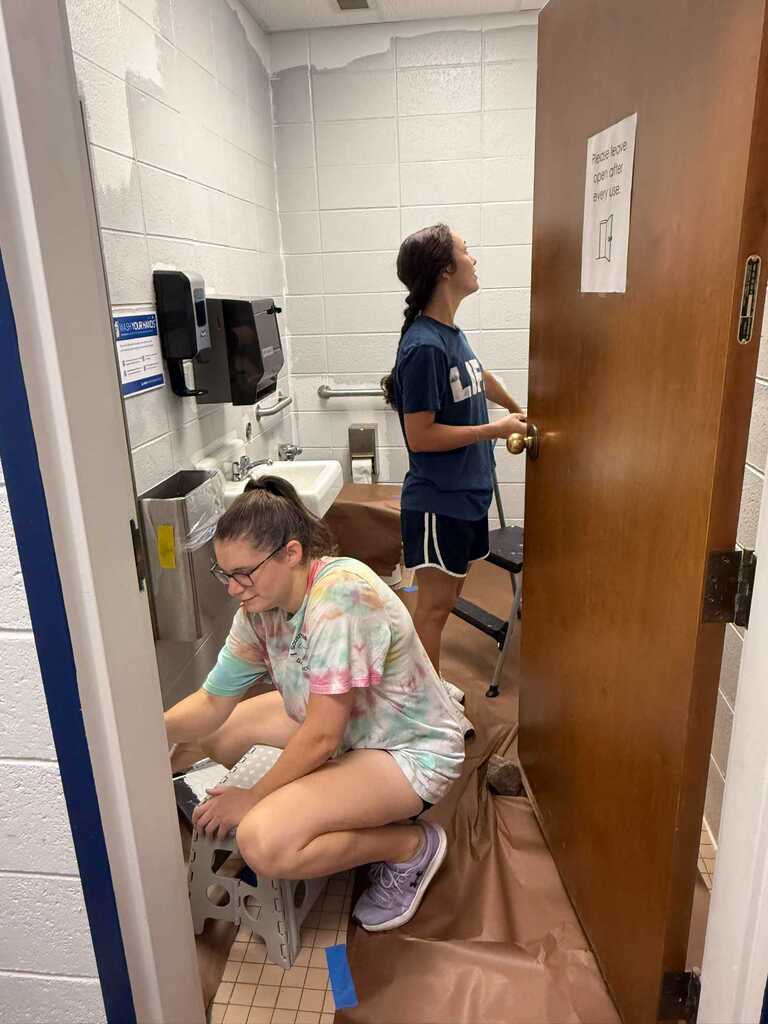 Two women work in a bathroom, one kneeling at a sink, the other opening a door. Tiled floor, white walls, and a sink with paper towels.