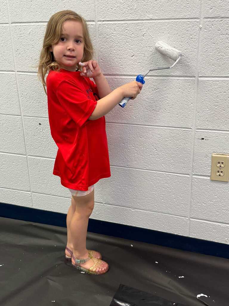 A young child holds a paint roller, applying white paint to a wall with a blue stripe.