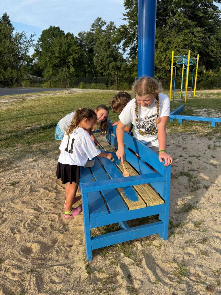 Four children are painting a blue bench. Trees and play equipment are in the background.