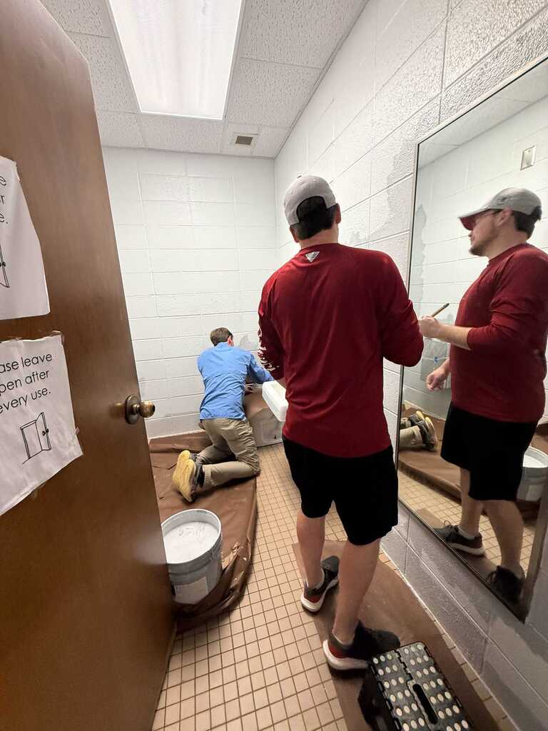 Three people in a bathroom. One is kneeling, one is standing, and one is reflected in the mirror.