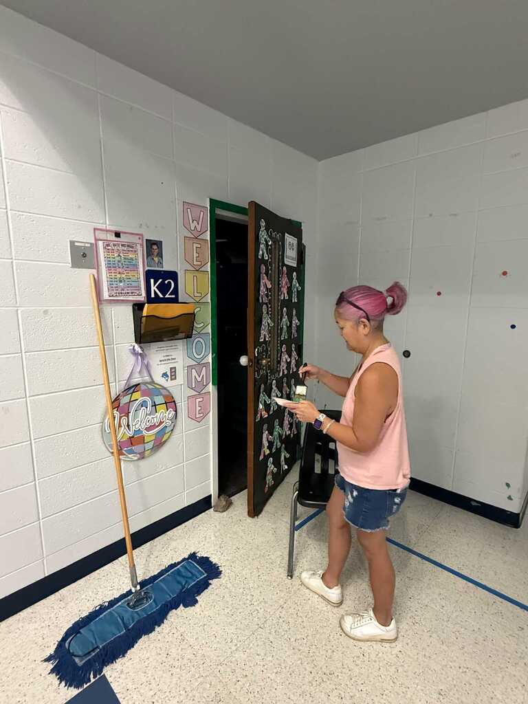 A woman cleans a classroom with a dust mop, wearing a pink shirt and denim shorts.