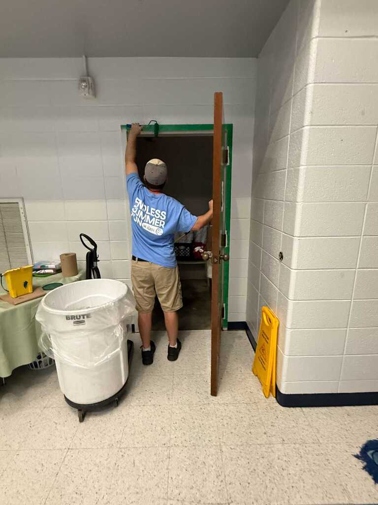 A man in a blue shirt and shorts holds an open door in a tiled room.
