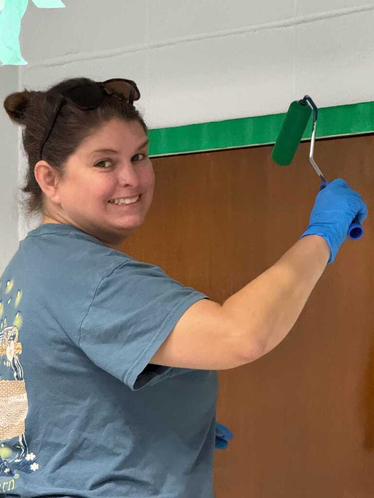 A woman paints a door with a roller. She wears a blue shirt and gloves, and her hair is tied back.
