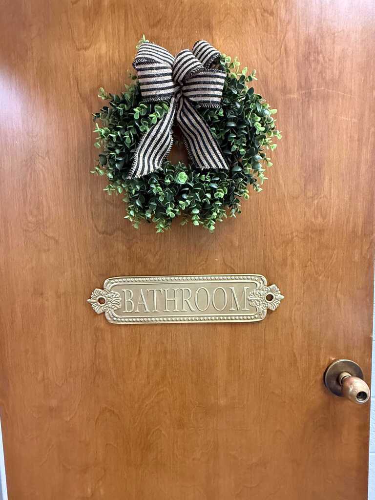 A wooden door features a wreath above a "BATHROOM" sign. The wreath has green leaves and a black and white ribbon bow.