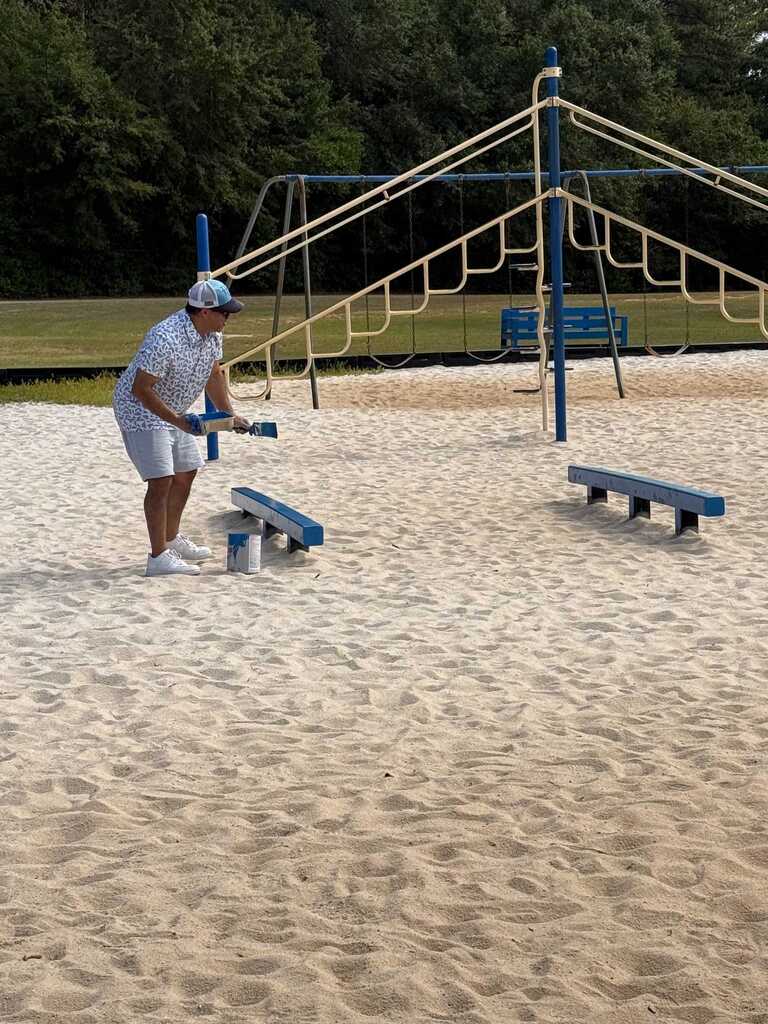 Person in hat and cap standing on sand holding a tool near a climbing structure.