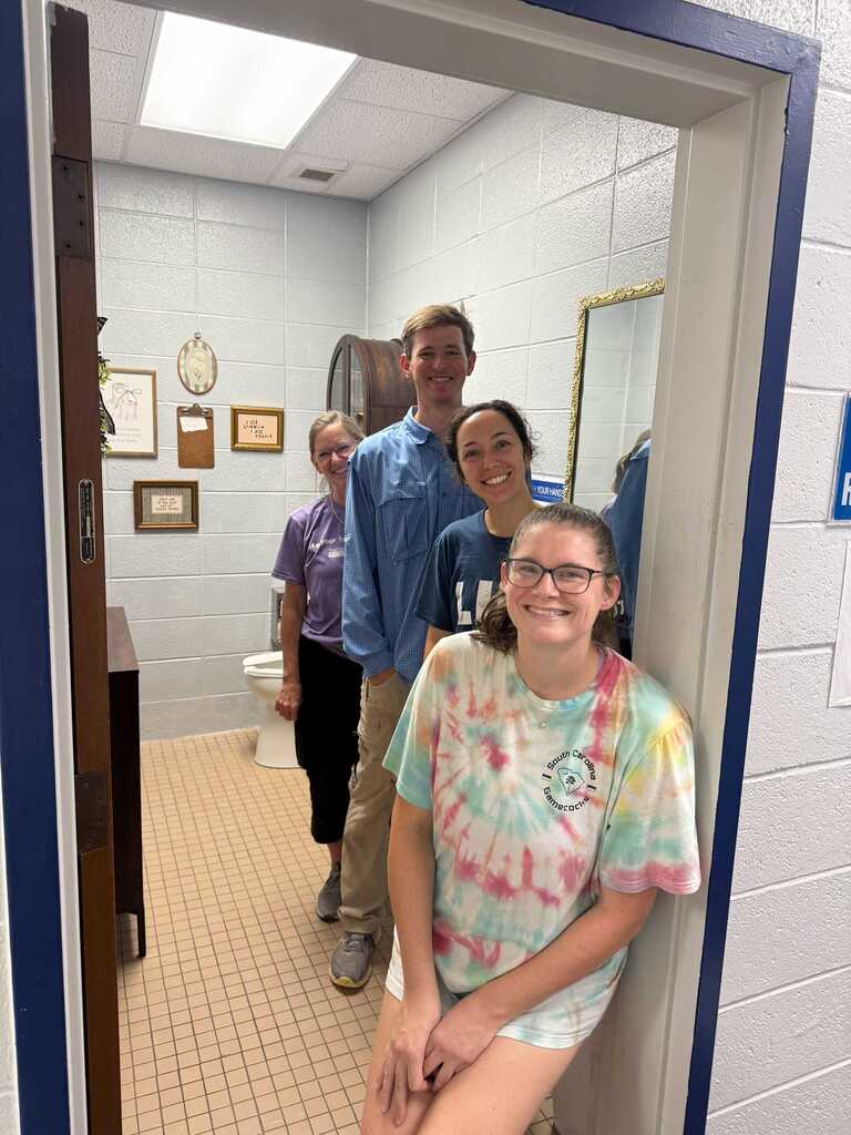Four people in a small bathroom. Three are standing, one sitting. Tiled floor, white walls. A mirror reflects two of them.